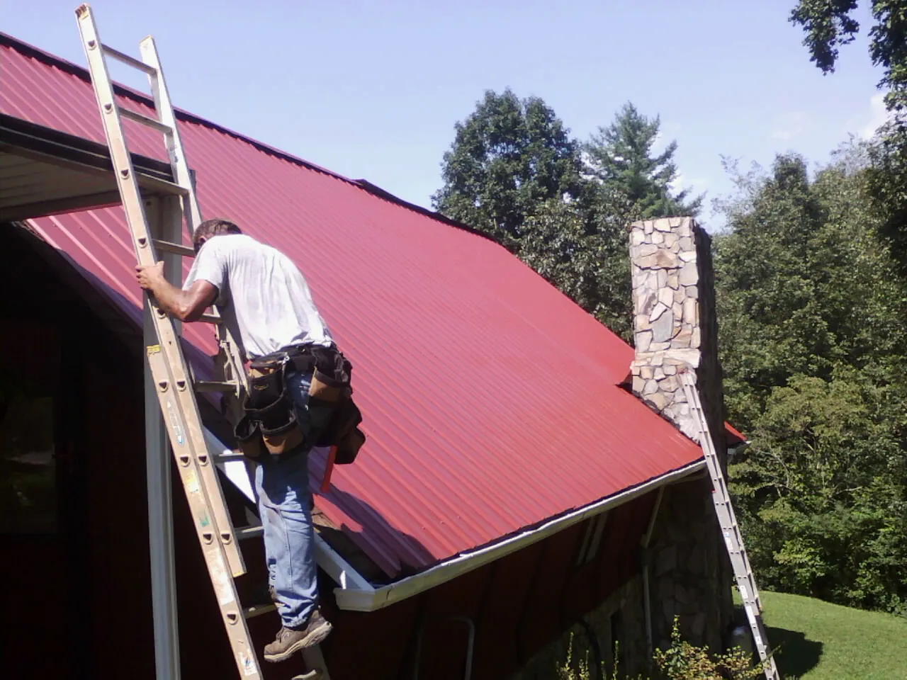 Roofer on a ladder beside a red metal roof and stone chimney — Soffit & Fascia Repair by Roofing Brothers in East Berlin